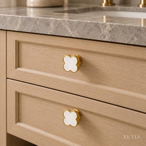 May include: Two clover-shaped cabinet knobs featuring a white, iridescent inlay surrounded by a polished gold-tone metal rim. The knobs are mounted on light-toned wooden drawers with a visible grain pattern. The text XUTIA is visible.