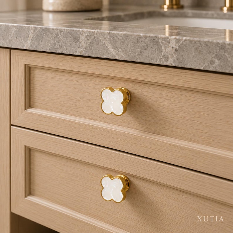 May include: Two clover-shaped cabinet knobs featuring a white, iridescent inlay surrounded by a polished gold-tone metal rim. The knobs are mounted on light-toned wooden drawers with a visible grain pattern. The text XUTIA is visible.
