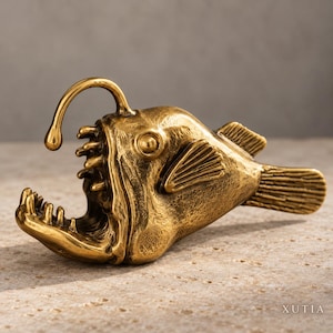 May include: A brass-colored anglerfish figurine with a textured metallic finish. The sculpture features a prominent lure extending from the head, sharp teeth, a wide open mouth, and detailed fins. The word XUTIA is printed in the bottom right corner.
