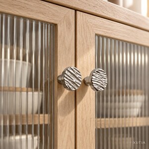 May include: A light oak wood cabinet featuring two doors with vertical reeded glass panels and round, silver-toned metal knobs with a textured, wavy surface. The word XUTIA is printed in small white letters in the bottom right corner.