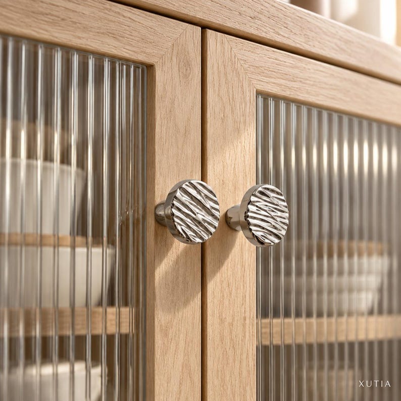 May include: A light oak wood cabinet featuring two doors with vertical reeded glass panels and round, silver-toned metal knobs with a textured, wavy surface. The word XUTIA is printed in small white letters in the bottom right corner.