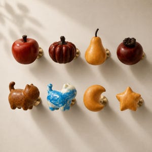 May include: A set of eight decorative cabinet knobs featuring various shapes including a red apple, a brown pumpkin, a yellow pear, a dark brown fruit, two cat figures in brown and blue, a yellow crescent moon, and a yellow star, all with brass bases.