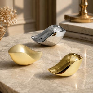 May include: Three bird-shaped decorative figurines with smooth, curved surfaces. One figurine has a reflective silver finish, while the other two feature a matte gold finish and a polished gold finish, respectively, all arranged on a flat surface.