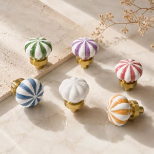 May include: A collection of six ceramic cabinet knobs with a fluted, pumpkin-like shape. Each knob features a glossy white base with alternating vertical stripes in green, purple, red, blue, orange, or solid white, all mounted on a brass base.