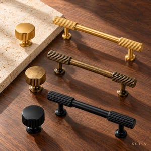 May include: A collection of cabinet hardware including three cylindrical knobs and three matching bar pulls in brushed gold, antique brass, and matte black finishes. Each piece features a distinct ribbed texture on the ends.