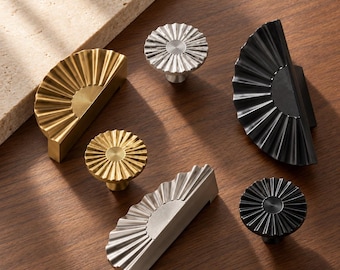 Könnte beinhalten: Eine Sammlung von Metall-Möbelbeschlägen, bestehend aus drei halbkreisförmigen fächerartigen Griffen und drei runden Knöpfen in Gold-, Silber- und Schwarz-Optik. Jedes Teil hat eine radial gerippte Textur und ein zentrales Detail. Der Text XUTIA ist unten rechts sichtbar.