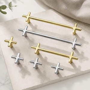 May include: A collection of metal cabinet hardware featuring two long bar pulls and four small knobs, all designed with a cross-shaped aesthetic. The set includes both polished gold-tone and silver-tone finishes, showcasing a smooth, reflective surface.