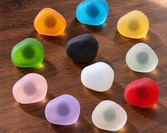 May include: A collection of eleven smooth, rounded, frosted glass magnets in various colors including orange, blue, yellow, green, black, white, pink, red, purple, and off-white. Each magnet features a dark circular center. The text XUTIA is visible.
