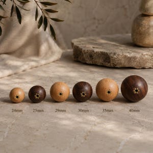 May include: A row of six spherical wooden beads with brass-lined center holes, arranged by size from left to right. The beads are labeled with their respective diameters: 24mm, 25mm, 29mm, 30mm, 35mm, and 40mm. The wood tones vary from light to dark.