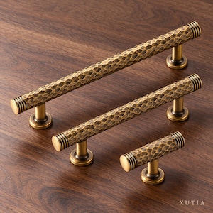 May include: Three brass cabinet pulls with a hammered texture and ribbed ends. Each handle features a cylindrical bar supported by two circular bases, except for the smallest knob which has one base. The brand name XUTIA is visible.