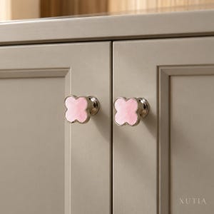 May include: Two clover-shaped cabinet knobs with a polished silver-tone metal base and a smooth, light pink stone inlay. The knobs are mounted on two adjacent light gray cabinet doors. The text XUTIA is visible in the bottom right corner.