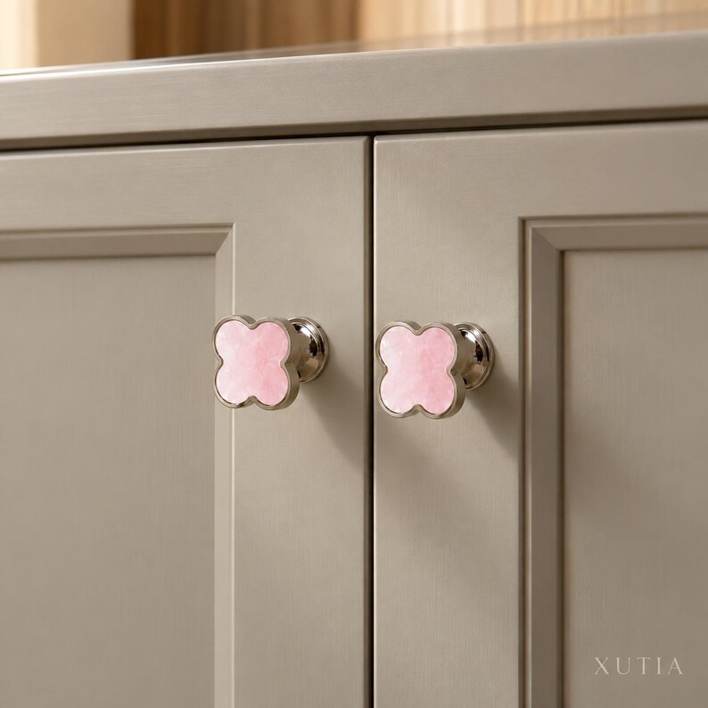 May include: Two clover-shaped cabinet knobs with a polished silver-tone metal base and a smooth, light pink stone inlay. The knobs are mounted on two adjacent light gray cabinet doors. The text XUTIA is visible in the bottom right corner.
