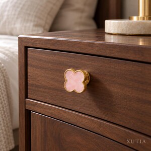 May include: A clover-shaped drawer knob featuring a polished gold-tone metal edge and a smooth, light pink stone inlay. The knob is mounted on a dark brown wooden drawer front with visible wood grain. The text XUTIA is printed in the bottom right.