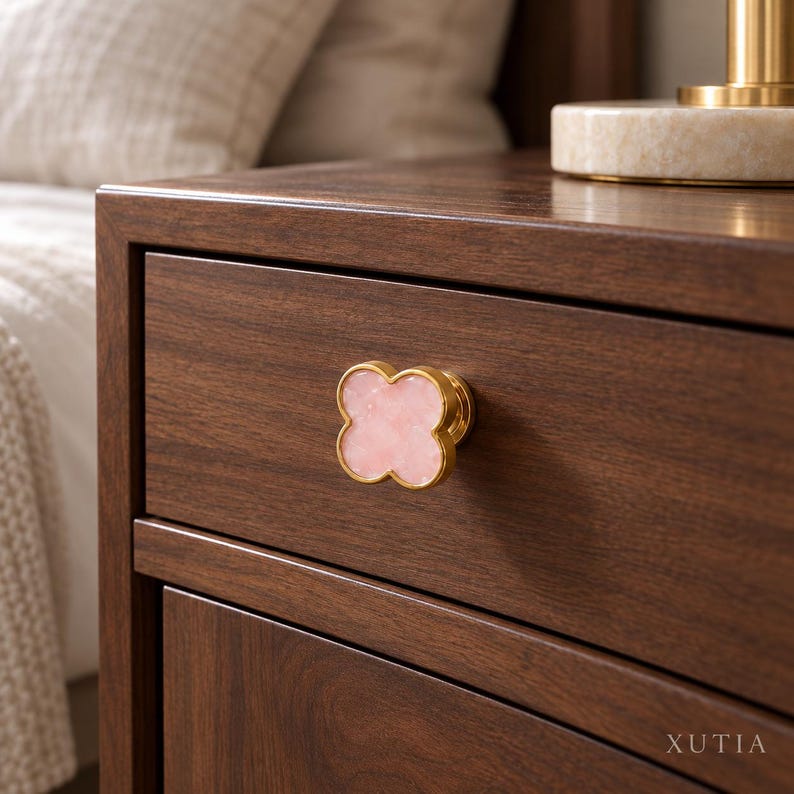 May include: A clover-shaped drawer knob featuring a polished gold-tone metal edge and a smooth, light pink stone inlay. The knob is mounted on a dark brown wooden drawer front with visible wood grain. The text XUTIA is printed in the bottom right.