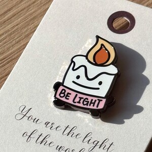 May include: A white enamel pin shaped like a candle with a flame, a simple face, and a pink base featuring the text BE LIGHT. The pin is attached to a white card printed with the text You are the light of the world and MATTHEW 5:14.