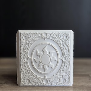 Puede incluir: Un azulejo decorativo cuadrado de color blanco con un motivo solar en relieve en el centro, rodeado de intrincados adornos de filigrana y pequeños iconos de sol en cada esquina. El diseño es elevado, creando una superficie texturizada mate.