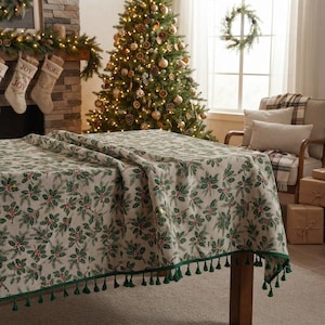 May include: A rectangular tablecloth featuring a repeating pattern of green holly leaves and red berries on a cream-coloured background. The edges of the fabric are finished with a decorative fringe of small, dark green tassels hanging from the hem.