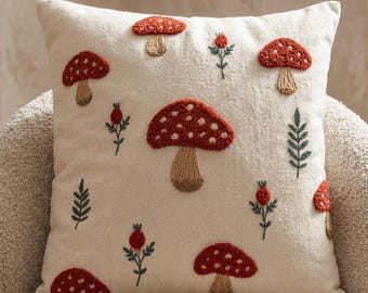 Op de afbeelding: Een vierkant gebroken wit sierkussen met geborduurde rode paddenstoelen met witte stippen, beige stelen en kleine groene botanische takjes met rode bessen verspreid over de voorzijde in een herhalend patroon.