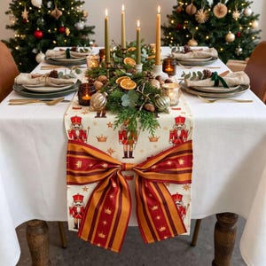 May include: A white table runner featuring a repeating pattern of red and gold nutcrackers, stars, and crowns, finished with a large, decorative red and gold striped bow hanging over the edge of a table set for a holiday meal.