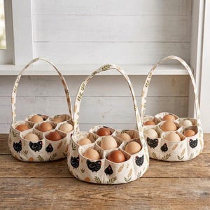Può includere: Tre cestini per uova in tessuto color bianco sporco con un manico, decorati con un motivo ripetuto di galline nere e steli di grano verdi. Ogni cestino contiene diverse uova marroni e bianche in scomparti circolari individuali.