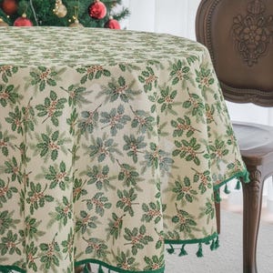 Puede incluir: Un mantel redondo con base de color crema y un patrón repetitivo de hojas de acebo verdes, agujas de pino y pequeñas bayas rojas. El borde de la tela está rematado con un ribete verde oscuro adornado con pequeñas borlas verdes colgantes.