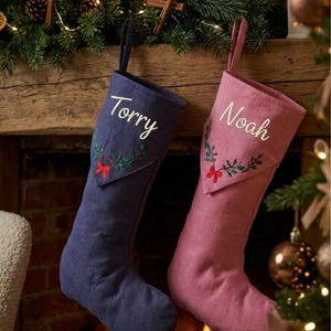 Peut inclure: Deux chaussettes de Noël personnalisées, une bleu foncé et une rose poudré, chacune avec un prénom brodé en blanc, Torry ou Noah, et un motif de feuilles de houx vertes avec un petit nœud rouge sur le revers, suspendues à une cheminée.