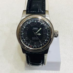May include: An Oris automatic wristwatch with a black textured dial, silver-tone hands, and Arabic numerals. The watch features a date pointer hand, a fluted bezel, and a black leather strap with a glossy finish. The dial displays Oris 25 Jewels.