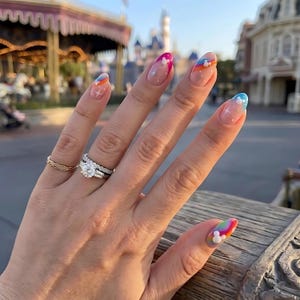 May include: Oval-shaped nails featuring a clear base with colorful rainbow-colored tips, small white star accents, and tiny white three-circle character icons. A gold braided ring and a silver-toned diamond engagement ring are worn on the finger.