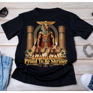 May include: A black short-sleeved t-shirt featuring a graphic print of an Egyptian-style figure wearing a headdress, flanked by two stone pillars and two camels. The text "Proud To Be Shriner" is printed in gold lettering at the bottom of the design.