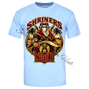May include: A light blue t-shirt featuring a graphic print of a figure in traditional desert attire holding a curved sword. The design includes the text SHRINERS NOBLE, palm trees, a desert landscape, and a gold emblem with a star and crescent.