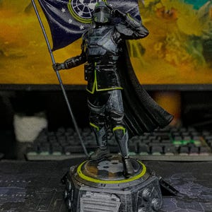 May include: A detailed figurine of a soldier in black and grey tactical armor with neon green accents, saluting while holding a dark blue flag featuring a white circular emblem. The figure stands on a hexagonal base with a metallic finish.