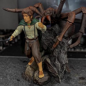 Pode incluir: Uma estatueta pintada de um personagem de cabelo castanho, vestindo uma camisa creme, calças castanhas e uma capa verde, segurando uma pequena espada prateada e um frasco brilhante. Atrás do personagem está uma grande aranha castanha numa rocha.