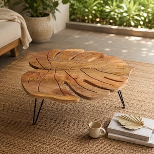 May include: A low coffee table with a tabletop shaped like a large monstera leaf, crafted from light brown wood with visible grain and carved vein details. The table is supported by three thin, black metal hairpin legs.