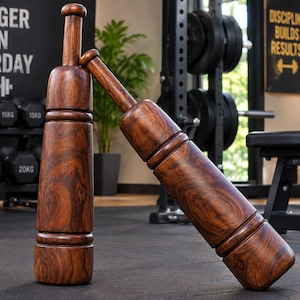 May include: Two dark brown wooden exercise clubs with a smooth, polished finish and a distinct grain pattern. One club stands vertically while the other leans against it at an angle, both featuring a cylindrical body with rounded ends and a handle.