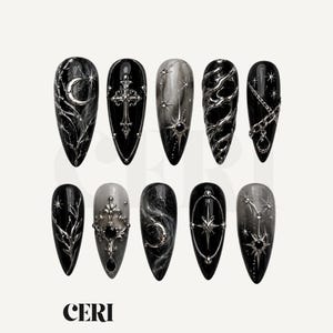 May include: A set of ten almond-shaped press-on nails featuring black and grey gradients with metallic silver 3D embellishments including crosses, crescent moons, stars, chains, and intricate filigree patterns. The brand name CERI is visible.