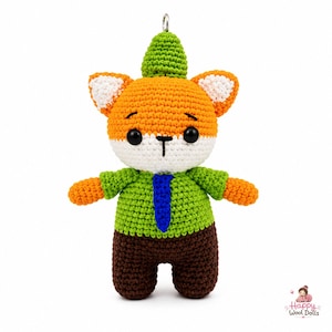 May include: A crocheted fox doll featuring orange and white yarn, wearing a green shirt with a blue tie and brown pants. The fox has black bead eyes, a small black stitched nose, and a green pointed hat. A metal loop is attached to the top.
