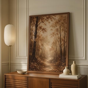 Könnte beinhalten: Ein gerahmter, rechteckiger Wandkunstdruck, der einen Waldweg im Herbst in warmen Braun- und Goldtönen zeigt. Das Kunstwerk steht auf einem hölzernen Sideboard neben einer hohen Stehlampe, einer kleinen Keramikschale und zwei beigen Vasen.