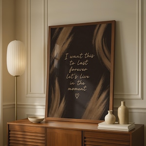 May include: A framed wall art print featuring a dark brown background with abstract gold brushstrokes and the text "I want this to last forever let's live in the moment" written in a gold script font, finished with a small gold heart icon.
