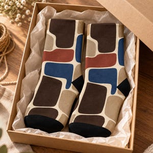 May include: A pair of crew socks featuring a geometric pattern of irregular shapes in dark brown, tan, terracotta red, and blue on a cream background. The socks have solid black toe and heel sections and are presented inside a cardboard gift box.