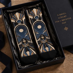 May include: A pair of black socks featuring a gold and blue train graphic with an arched design, presented in a black gift box with the text A SMALL LUXURY FOR A BRIGHTER DAY printed in gold on the interior lid of the box.