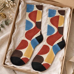May include: A pair of crew socks featuring an abstract pattern of curved shapes in shades of red, blue, yellow, and dark brown on a cream background, with solid black toe and heel sections, presented inside a rectangular cardboard gift box.