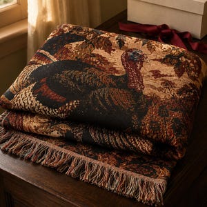 Könnte beinhalten: Eine gefaltete Webdecke mit einem detaillierten Wandteppichmuster, das einen Truthahn umgeben von herbstlichem Laub in den Farben Schwarz, Rost, Braun und Beige zeigt. Das rechteckige Textil ist an der Unterkante mit Fransen versehen.