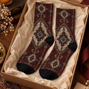 May include: A pair of crew socks featuring a deep burgundy base with a complex, symmetrical geometric pattern in shades of cream, navy blue, and light blue. The socks have solid black heels and toes, presented inside a cardboard gift box.