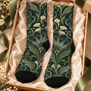 May include: A pair of crew socks featuring a dark green and sage botanical pattern with stylized fan-shaped leaves and swirling floral motifs. The socks have solid black heels and toes, presented inside a rectangular cardboard gift box.