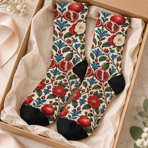 May include: A pair of crew socks featuring a botanical pattern of red pomegranates, white and red flowers, and blue leaves on a cream background. The socks have solid black heels and toes and are presented inside a cardboard gift box with tissue paper.