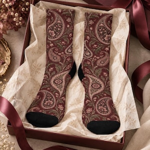 May include: A pair of crew socks featuring a deep burgundy background with an intricate paisley and floral pattern in shades of muted green, pink, and cream. The socks have solid black heels and toe sections, presented inside a gift box.