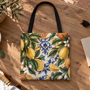 May include: A square tote bag with black shoulder straps featuring a vibrant print of yellow lemons, orange slices, and green leaves layered over a blue and white decorative tile pattern. The bag has a flat base and a structured fabric construction.