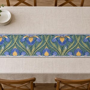 May include: A rectangular quilted table runner featuring a repeating pattern of blue and yellow iris flowers with long green leaves on a dark teal background, finished with a light blue border and a stitched diamond grid texture throughout.