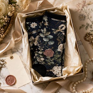 May include: A pair of dark navy blue socks featuring a detailed botanical print of white flowers, green leaves, and butterflies, finished with solid black cuffs and heels, presented inside a gift box lined with light beige tissue paper.