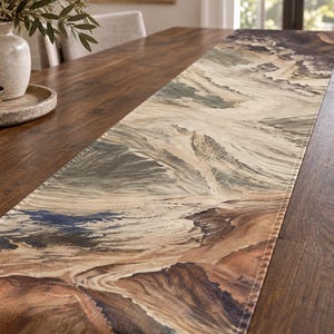 Könnte beinhalten: Ein rechteckiger Tischläufer mit einem gedruckten Landschaftsmotiv in Beige-, Braun-, Dunkelgrün- und Schieferblautönen. Der Stoff zeigt ein strukturiertes, bergähnliches Geländemuster mit genähten Kanten und liegt flach auf einer Oberfläche.